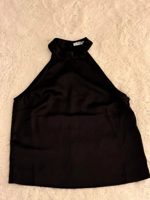 Svart halterneck topp  - Snygg svart halterneck topp från NA-KD i ett glansigt satinliknande material. Toppen har hög krage och är ärmlös, perfekt för att skapa en stilren look. Toppen är nästan helt ny (användes 1 gång)
