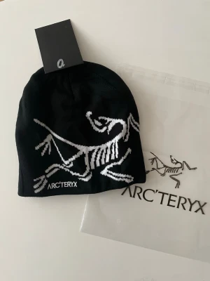 Svart mössa från Arc'teryx - Använt, Snygg svart mössa från Arc'teryx med vit logotyp och ikoniskt skelettmönster framtill. Tillverkad i mjukt stickat material som håller dig varm och bekväm. Perfekt accessoar för kalla dagar och outdoor-stil.