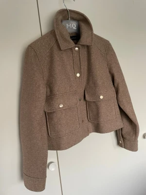 Beige ullcardigan från Zara - Snygg beige cardiganjacka från Zara i ullmix med krage och stora fickor framtill. Kortare modell med knappar i metall och raka ärmar. Perfekt för lager på lager och ger en clean look till din outfit.