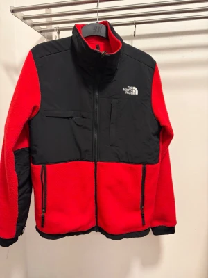 Röd och svart fleecejacka The North Face - Röd och svart fleecejacka från The North Face med hög krage, dragkedja framtill och flera fickor. Jackan har svarta detaljer på axlar och ärmar samt logga på bröstet och ryggen. Perfekt för kyliga dagar och outdoor vibes.