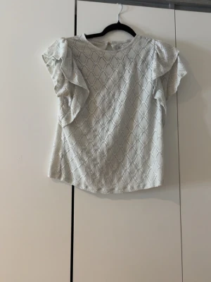 Vit blus med volangärmar - Snygg vit blus med korta volangärmar och diskret mönster i tyget. Blusen har rund halsringning och en liten knapp i nacken. Perfekt för en trendig och stilren look. Köptes här på plick men kom aldrig riktigt till användning.