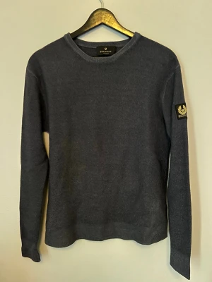 Mörkgrå stickad tröja från Belstaff - Mörkgrå stickad tröja från Belstaff med rund halsringning och lång ärm. Tröjan har en klassisk passform och en snygg logotyp-patch på vänster ärm. Perfekt för lager-på-lager och enkel att matcha med jeans eller chinos. Ny pris 2500