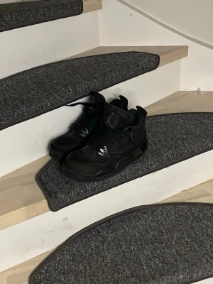 Air jordan 4 (Black Cats) - Snygga helsvarta Air Jordan 4 Retro sneakers med klassisk siluett och meshdetaljer på sidorna. Skorna har högt skaft, ikoniska plastdetaljer och är tillverkade i en mix av läder och textil. Perfekta för dig som gillar streetwear och vill ha en stilren look.