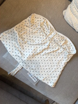 Ny i förpackning! Vit prickig topp med knyt - Supersöt vit topp med svarta prickar och knytband framtill. Toppen är axelbandslös och har en lätt croppad passform. Perfekt för att skapa en trendig och lekfull look. Materialet känns mjukt och stretchigt.