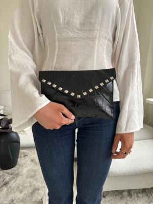 Clutch med silvernitar  - Med sina mått på 25 x 17 cm är den perfekt stor för det viktigaste – mobil, nycklar och små nödvändigheter – utan att kännas klumpig. Väskan är handgjord i hållbart veganskt läder, vilket ger en mjuk och exklusiv känsla samtidigt som den är ett mer medvetet val.