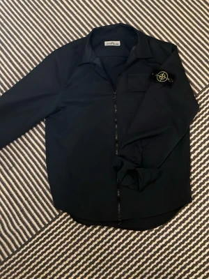Mörkblå stone island overshirt - Snygg svart overshirt från Stone Island med dragkedja framtill och klassisk krage. Har en bröstficka och den ikoniska Stone Island-loggan på vänster ärm. Tillverkad i ett slätt, tekniskt material som ger en clean och modern look. Perfekt lager på lager-plagg.
