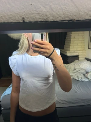 Vit croppad topp med volangärm - Snygg vit croppad topp med volangdetaljer på axlarna. T-shirten har rund halsringning och är tillverkad i ett mjukt bomullsmaterial. 