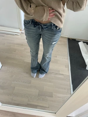 Blå bootcut delulu jeans med slitningar - Säljer ett par blå bootcut jeans med slitna detaljer och ljusa tvättar på låren och knäna. Jeansen har låg midja, klassiska fem fickor och är gjorda i denim. Perfekta för dig som gillar en avslappnad och trendig stil. Aldrig använt de är för stora för mig.