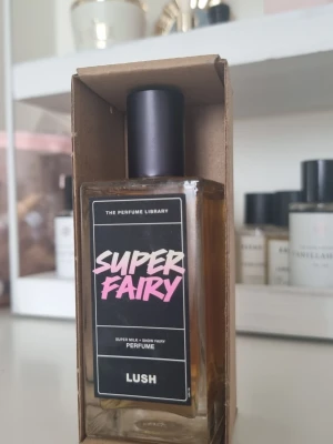 Lush Super Fairy parfym - Super Fairy parfym från Lush i en rektangulär glasflaska med svart etikett och rosa/vit text. Flaskan har svart lock och kommer i en enkel kartongförpackning. Perfekt för dig som gillar unika och lekfulla dofter.