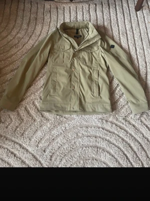 Grön / khaki jacka  från J.Lindeberg Stl M - Stilren beige utilityjacka från Jack & Jones med klassisk krage, bröstfickor med knappar och långa ärmar. Jackan har en rak passform och är tillverkad i bomullsmaterial. Perfekt för dig som gillar en clean och enkel look.