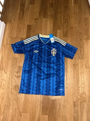 Blå Sverige fotbollströja Adidas - Säljer en blå fotbollströja från Adidas med Sveriges landslagsemblem på bröstet. Tröjan har ett retroinspirerat mönster, vita Adidas-ränder på axlarna och guldfärgade detaljer vid kragen. Perfekt för dig som vill stötta Sverige med stil.
