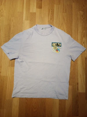 Ljuslila t-shirt med tryck från Zara - Ljuslila t-shirt från Zara med coolt färgglatt tryck på bröstet där det står 'EST. 75' och en tecknad detalj med ögon och texten 'YES RIGHT NOW!'. Klassisk passform och rund halsringning.