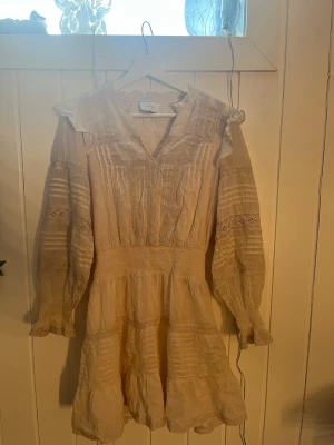 Beige spetsklänning från Neo Noir - Romantisk beige klänning från Neo Noir med v-ringning, volangdetaljer vid axlar och ärmslut samt spetsinslag och broderade partier. Långärmad modell med markerad midja och lätt utställd kjol. Perfekt för en bohemisk look.