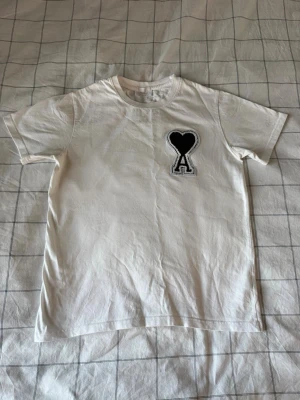 Vit t-shirt från Ami med hjärtdetalj - Vit t-shirt från Ami Alexandre Mattiussi med svart hjärta och bokstaven A broderat på bröstet. Klassisk passform med rund hals och korta ärmar. Tillverkad i mjuk bomull, perfekt för en clean och stilren look. Tröjan är i storlek M men passar bra i S också