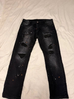 🔥 Trendiga svarta jeans med ripped-design & paint splatter – XXXL 🔥 - Säljer ett par riktigt snygga svarta jeans med slitningar (ripped) och paint splatter-detaljer som ger en modern och exklusiv streetwear-look. Byxorna är bekväma med stretch och sitter riktigt bra.                                                  Storlek: XXXL (med stretch). För referens är jag 192 cm lång och väger ca 90 kg, och byxorna passar mig riktigt bra utan att sitta tajt. De passar därför bra för dig som vill ha en bekväm, normal eller något lösare passform. 
