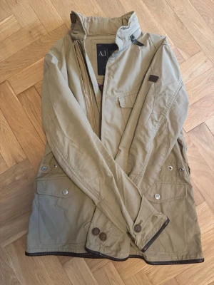 Fieldjacket från Armani Jeans - Stilren beige fieldjacket från Armani Jeans med flera fickor framtill, dragkedja och läderknappar. Jackan har hög krage. Perfekt för dig som vill ha en snygg och funktionell jacka till vardags. Självklart äkta✅