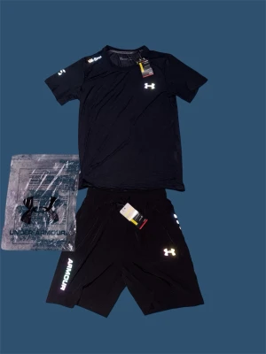 Svart träningsset från Under Armour - Snyggt svart träningsset från Under Armour med t-shirt och shorts. Båda plaggen har logga tryckt i vitt och reflekterande detaljer. Tillverkade i lätt och snabbtorkande material som passar perfekt för gym eller löpning. Shortsen har dragsko i midjan och tryck på benet.