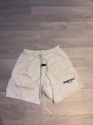 Grå Essentials Fear of God shorts - Snygga ljusgrå shorts från Essentials Fear of God med svart logga på vänster ben. De har elastisk midja med snörning och är tillverkade i mjuk bomull, perfekta för chill dagar. Klassisk passform och enkel stil som funkar till det mesta.