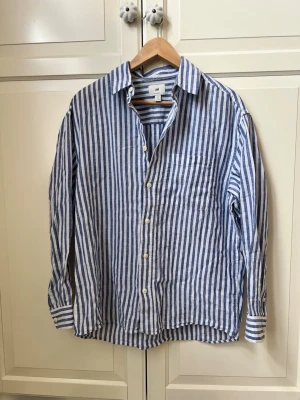 Randig linneskjorta från H&M - Snygg långärmad skjorta från H&M i blått och vitt med vertikala ränder. Tillverkad i luftig linne, perfekt för varma dagar. Klassisk krage, bröstficka och knäppning framtill. Skjortan har en relaxed fit och passar till många olika stilar.