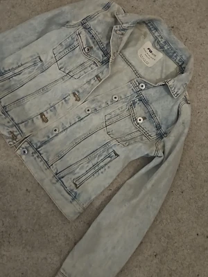 Ljusblå jeansjacka  - Snygg ljusblå jeansjacka med klassisk tvättad look. Jackan har två bröstfickor med lock och knappar, samt två sidofickor. Silverfärgade knappar framtill och på fickorna. Perfekt för en avslappnad och trendig stil.