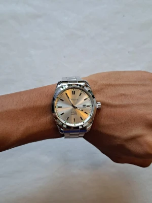 Silverfärgad klocka från Denuasi - Stilren klocka från Denuasi med silverfärgad urtavla och matchande metallarmband. Klockan har tydliga svarta markeringar, datumvisning och klassisk rund form. Perfekt accessoar för dig som gillar en clean och tidlös look.                   Storlek: 40mm | Armbandet går att justeras genom att ta bort länkar på armbandet.