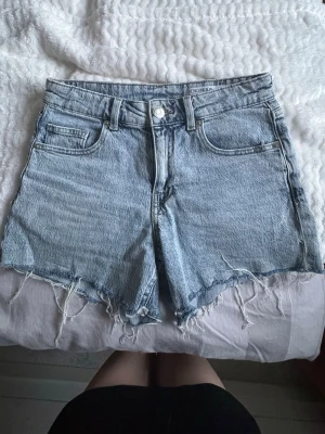 Ljusblå jeansshorts från Vero Moda - Säljer ett par ljusblå jeansshorts från Vero Moda med råa, fransiga benslut och klassisk femficksdesign. Shortsen har hög midja och är tillverkade i bomull för en skön och avslappnad känsla. Perfekta för varma dagar!