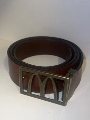 Brunt läderbälte med McDonald's-spänne - Snyggt brunt bälte i skinn med ett unikt metallspänne som har McDonald's ikoniska M-logga. Perfekt accessoar för dig som vill sticka ut och gillar retro vibes. Bältet har en klassisk design och passar till många olika outfits.