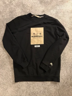 Svart sweatshirt från Burberry - Svart sweatshirt från Burberry med beige patch och logga på bröstet. Rund halsringning, långa ärmar och ribbade muddar. Liten Burberry-detalj på ärmen och en extra logotyp under patchen. Tillverkad i mjukt bomullsmaterial.