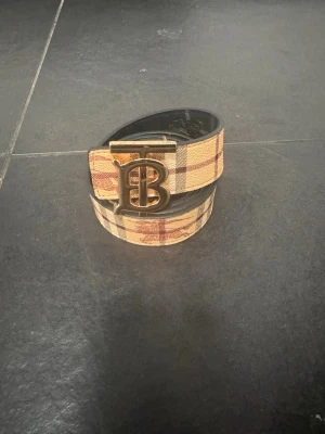 Rutig bälte med gulddetalj - Snyggt bälte från med klassiskt rutigt mönster i beige, rött och svart. Bältet har ett stort guldfärgat spänne med ikoniska TB-logga. Perfekt accessoar för att lyfta din outfit.