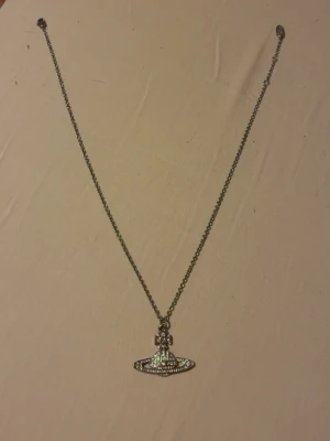Vivienne Westwood halsband med strass - Snyggt halsband från Vivienne Westwood med det ikoniska orb-hänget täckt av glittrande strass. Kedjan är silverfärgad och hänget har en unik form som verkligen sticker ut. Perfekt accessoar för dig som gillar statement-smycken och vill ha något som syns.