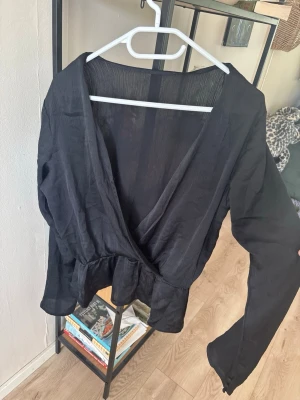 Svart omlottblus med volang - Stilren svart omlottblus med djup v-ringning och volang nedtill. Blusen har långa ärmar och är tillverkad i ett tunt, lätt transparent tyg som ger en luftig känsla. Perfekt att styla med jeans eller kjol för en trendig look. Finns ingen lapp i tröjan.