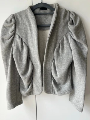 Grå jerseykavaj med puffärmar - Snygg ljusgrå jerseykavaj med markerade puffärmar och draperade detaljer längs sidorna. Jackan har en öppen front med dold hake för stängning och är tillverkad i mjukt skönt material. Perfekt för en trendig och avslappnad look.