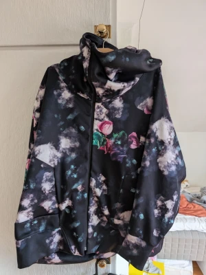 Balmung, oversized hoodie - BALMUNG ”Dust Flower”, jacka/hoodie med grafiskt tryck. Unisex.  Från 19S/S 2019 Vår/sommarkollektionen.   BALMUNG är känt för sin överdimensionerade silhuett och unika grafiska mönster. Detta är ett distinkt plagg med BALMUNGs signaturdetalj, den höga halsen som täcker ända upp till munnen och en stor silhuett. Jackan har  två fickor  100% polyester   Längd: 78 cm Axelbredd: går ej att mäta då ärmarna och Axelpartiet går in i varandra  Ärmlängd: ca 99 cm Bredd: 94 cm Storlek: Free 