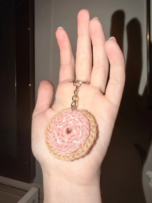 Virkad donut med glasyr och strössel nyckelring  - Söt handgjord nyckelring formad som en donut i virkat garn. Donuten är rosa med beige kant och strössel. Perfekt att hänga på väskan eller nyckelknippan. Levereras i en liten organzapåse. Inspirerad av Homer Simpson-donuten.