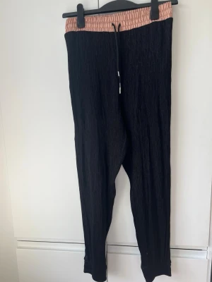 Svarta mjukisbyxor från Zara M - Säljer ett par svarta mjukisbyxor från Zara i storlek M. Byxorna har en bred, glansig resårmidja i ljusrosa och dragsko framtill. Materialet är mjukt och har en lätt struktur, perfekta för chill dagar. Snygg kontrast mellan midja och byxa!