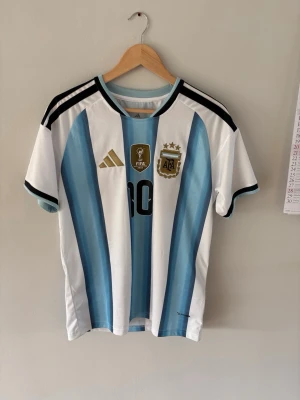 Argentina Messi matchtröja Adidas - Säljer en officiell Argentina matchtröja från Adidas med Messi och nummer 10 på ryggen. Tröjan har klassiska blå och vita ränder, svarta detaljer och guldiga märken på bröstet. Perfekt för fotbollsfans som vill stötta sitt lag med stil.