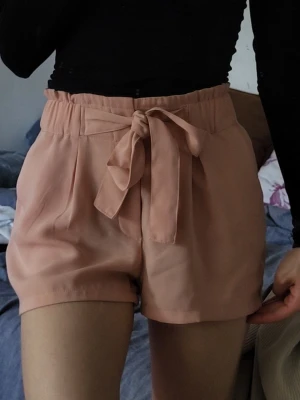 Beiga shorts med knytband - Snygga beiga shorts med hög midja och brett knytband framtill. Shortsen har en lös passform och är tillverkade i ett mjukt, lätt tyg som känns skönt mot huden. Perfekta för varma dagar och enkla att matcha med olika toppar.