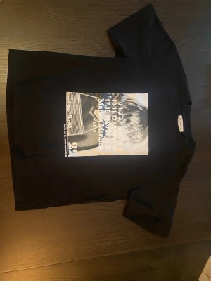 Svart t-shirt Maison Margiela print - Cool svart t-shirt från Maison Margiela med stort grafiskt print framtill i vitt och beige toner. Klassisk passform med rund hals och korta ärmar. Tillverkad i mjuk bomull för skön känsla och stilren look. Kom gärna med pris förslag