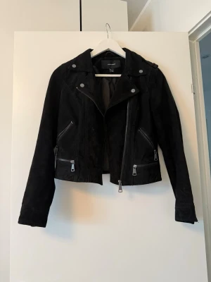 Svart mockajacka från Vero Moda - Snygg svart skinnjacka från Vero Moda med klassisk bikerstil, sned dragkedja och flera dragkedjeförsedda fickor. Defektfinns i underarmen se foto