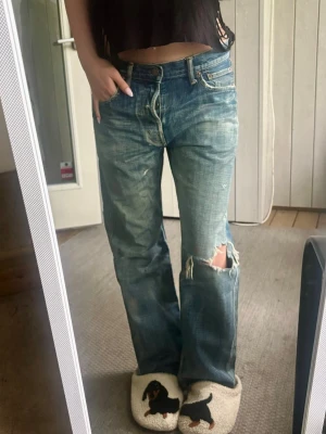 Blå slitna jeans med hål - Säljer ett par blå  jeans med slitningar och stort hål på ena knät. Ena knappen går inte att knäppa men tror det är designen på jwanses som är så. Jeansen har klassisk femficksmodell, låg midja och en avslappnad passform. Perfekta för en cool och avslappnad stil.
