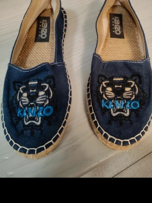 Blå Kenzo ballerinaskor med tiger - Snygga blå ballerinaskor från Kenzo med broderad tiger och logga i blått och vitt framtill. Skorna är i canvas med flätad sula och har en avslappnad espadrille-look. Perfekta för dig som gillar streetstyle och vill sticka ut med coola detaljer.