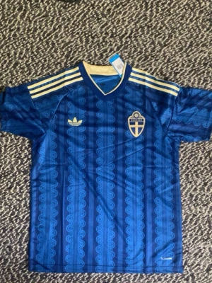 Blå Sverige fotbollströja Adidas - Kan mötas upp i Malmö. Säljer en blå fotbollströja från Adidas med Sveriges landslagsemblem på bröstet. Tröjan har korta ärmar, guldfärgade ränder på axlarna och ett retroinspirerat mönster. Tillverkad i lätt och ventilerande material, perfekt för träning eller match.