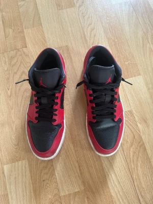 Air Jordan 1 Low svart/röd - Säljer ett par Air Jordan 1 Low i svart och rött läder med klassisk Nike Swoosh på sidan och Air Jordan-logga på hälen. Skorna har svarta skosnören, perforerad tåbox och vit sula. Perfekta för dig som gillar streetwear och ikoniska sneakers.