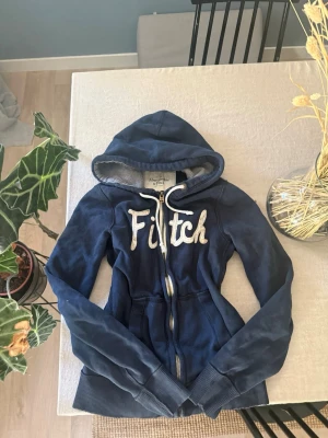 Marinblå hoodie Abercrombie & Fitch - Snygg marinblå hoodie från Abercrombie & Fitch med dragkedja och stor vit broderad text 'Fitch' på bröstet. 