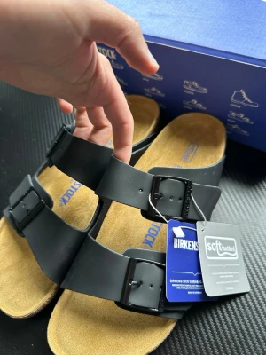 Svarta Birkenstock Arizona sandaler - Säljer ett par svarta Birkenstock Arizona sandaler med två breda remmar och justerbara spännen. Fotbädden är mjuk och har en beige mockayta, sulan är i kork och gummi. Klassisk och tidlös modell som är superbekväm för vardagsbruk.