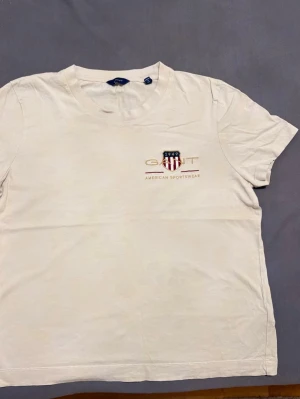 Vit GANT t-shirt med broderad logga - Snygg vit t-shirt från GANT med broderad logga och texten 'American Sportswear' på bröstet. Klassisk rund halsringning och korta ärmar. Perfekt basplagg med stilren design och mjukt material.