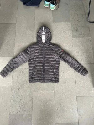 Grå pufferjacka med huva - Snygg grå pufferjacka från colmar med quiltad design och huva. Jackan har dragkedja framtill, två sidofickor med dragkedja och en logotyp på vänster ärm. Perfekt för kalla vinterdagar och riktigt lätt att bära.