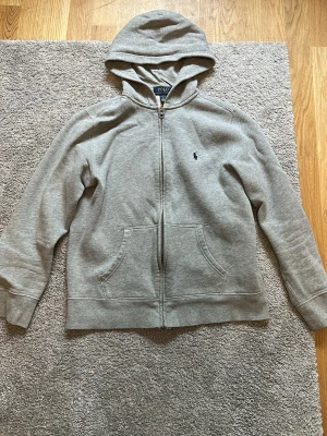 Grå hoodie från Polo Ralph Lauren - Klassisk grå hoodie från Polo Ralph Lauren med dragkedja framtill och två fickor. Tröjan har huva och den ikoniska lilla loggan broderad på bröstet. Tillverkad i mjuk bomullsblandning som är skön att ha på sig.