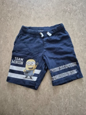 Shorts - Mörkblå shorts med tryck av en Minion och texten 'Team Minion' på ena benet. Shortsen har vita ränder och snörning i midjan. Perfekta för sommaren och tillverkade i mjuk bomull.
