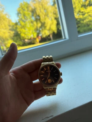 Guldig klocka från Versus Versace - Snygg guldig klocka från Versus Versace med svart urtavla och guldfärgade detaljer. Klockan har en klassisk länkarmband i metall och stilren design med Versus-logga på spännet. Perfekt accessoar för att lyfta din outfit.